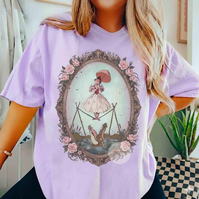 Puede incluir: Una camiseta de color morado claro con un marco ovalado central decorado con rosas rosas. Dentro del marco, una ilustraci&oacute;n muestra a una figura con vestido y sombrilla haciendo equilibrio sobre una cuerda sobre un caim&aacute;n marr&oacute;n en el agua.