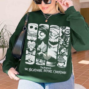 May include: Dark green sweatshirt with a graphic from Tim Burton's "The Nightmare Before Christmas." The design features characters such as Jack Skellington, Sally, and Oogie Boogie in white. The text "Tim Burton's The Nightmare Before Christmas" is printed below the graphic.