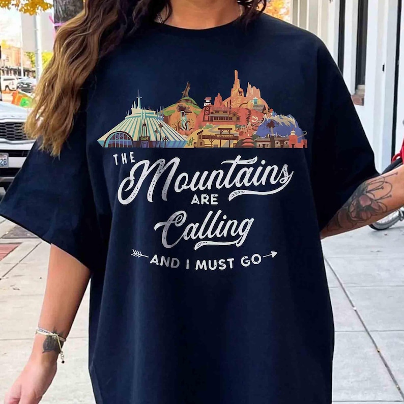 May include: A dark navy blue short-sleeve t-shirt featuring a colorful graphic of various mountain landscapes and buildings. The text printed in white script and block letters reads, "THE Mountains ARE Calling AND I MUST GO" with arrows.