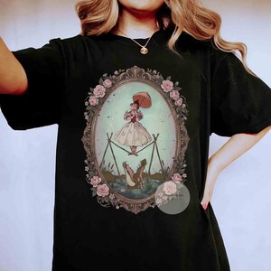 Puede incluir: Una camiseta negra con un gr&aacute;fico central dentro de un marco ovalado decorado con rosas rosadas. La ilustraci&oacute;n muestra a una persona con un vestido claro sosteniendo una sombrilla mientras camina sobre una cuerda floja sobre un caim&aacute;n.