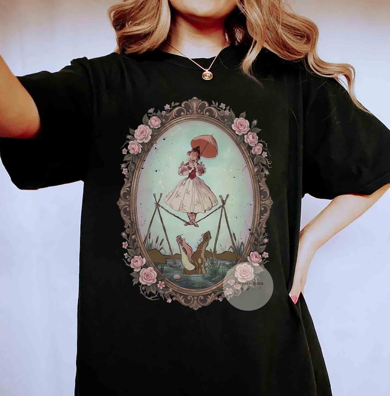 Puede incluir: Una camiseta negra con un gr&aacute;fico central dentro de un marco ovalado decorado con rosas rosadas. La ilustraci&oacute;n muestra a una persona con un vestido claro sosteniendo una sombrilla mientras camina sobre una cuerda floja sobre un caim&aacute;n.