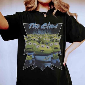 K&ouml;nnte beinhalten: Schwarzes T-Shirt mit einer Grafik von gr&uuml;nen, drei&auml;ugigen Aliens aus dem Film Toy Story. Die Aliens befinden sich in einem dreieckigen Rahmen mit dem Text "The Claw" in Blau. Die Aliens tragen blaue Uniformen.