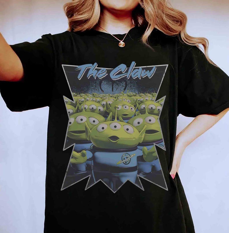 K&ouml;nnte beinhalten: Schwarzes T-Shirt mit einer Grafik von gr&uuml;nen, drei&auml;ugigen Aliens aus dem Film Toy Story. Die Aliens befinden sich in einem dreieckigen Rahmen mit dem Text "The Claw" in Blau. Die Aliens tragen blaue Uniformen.
