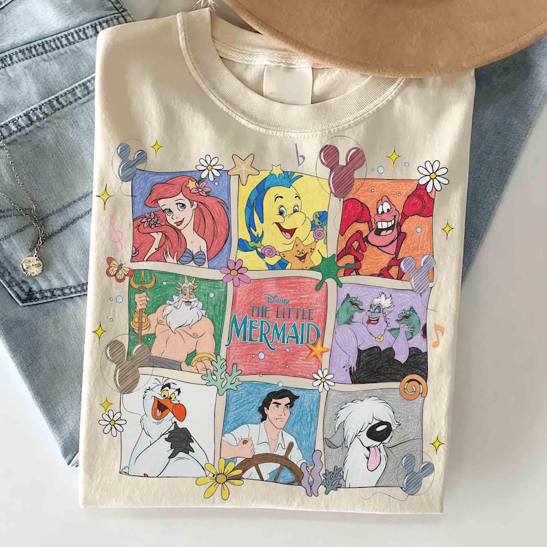 May include: Cream-colored crew neck sweatshirt featuring a colorful graphic of characters from Disney's The Little Mermaid. The design includes Ariel, Flounder, Sebastian, King Triton, Ursula, and Prince Eric. The title "The Little Mermaid" is also displayed.