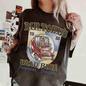 K&ouml;nnte beinhalten: Dunkelgraues T-Shirt mit einer Grafik von Mr. Toad in einem roten Auto aus der Attraktion Mr. Toad's Wild Ride. Das Design enth&auml;lt den Text "Mr. Toad's Wild Ride" und "Fantasyland Disneyland".