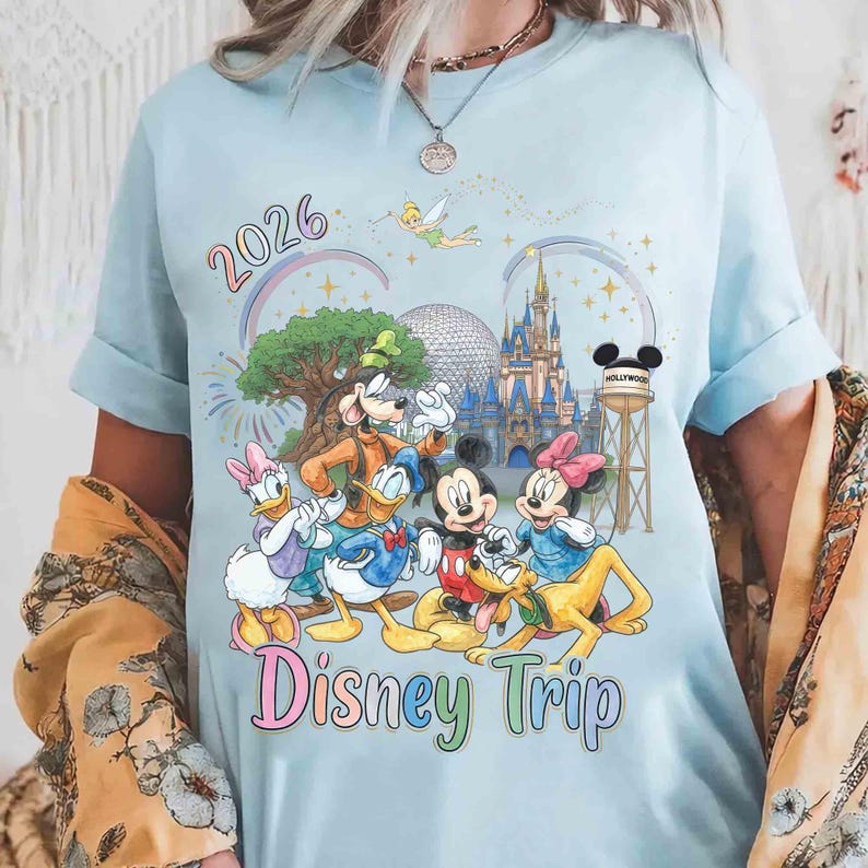 May include: A light blue t-shirt featuring a colorful graphic of Disney characters including Mickey Mouse, Minnie Mouse, Donald Duck, Daisy Duck, Goofy, Pluto, and Tinker Bell, with the text 2026 Disney Trip and illustrations of a castle and water tower.