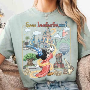 Op de afbeelding: Lichtgroen t-shirt met een kleurrijke afbeelding van Mickey Mouse als tovenaar, een kasteel en de tekst "Some Imagination, Huh?". Het ontwerp bevat themaparkelementen en een toverstaf.