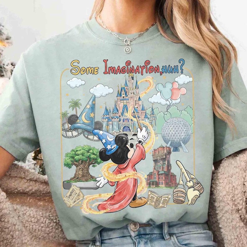 Op de afbeelding: Lichtgroen t-shirt met een kleurrijke afbeelding van Mickey Mouse als tovenaar, een kasteel en de tekst "Some Imagination, Huh?". Het ontwerp bevat themaparkelementen en een toverstaf.