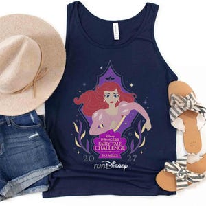 May include: A navy blue tank top featuring a graphic of Ariel with red hair, a purple top, and pink sleeves. The text reads Disney Princess Fairy Tale Challenge 19.3 Miles 2027 runDisney. Also includes a beige sun hat, denim shorts, and striped sandals.