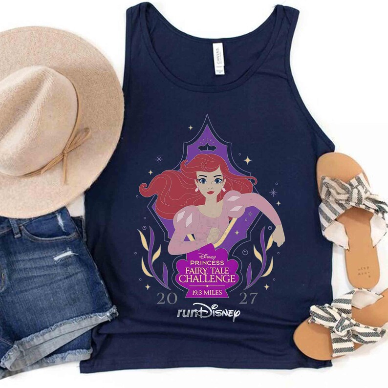 May include: A navy blue tank top featuring a graphic of Ariel with red hair, a purple top, and pink sleeves. The text reads Disney Princess Fairy Tale Challenge 19.3 Miles 2027 runDisney. Also includes a beige sun hat, denim shorts, and striped sandals.