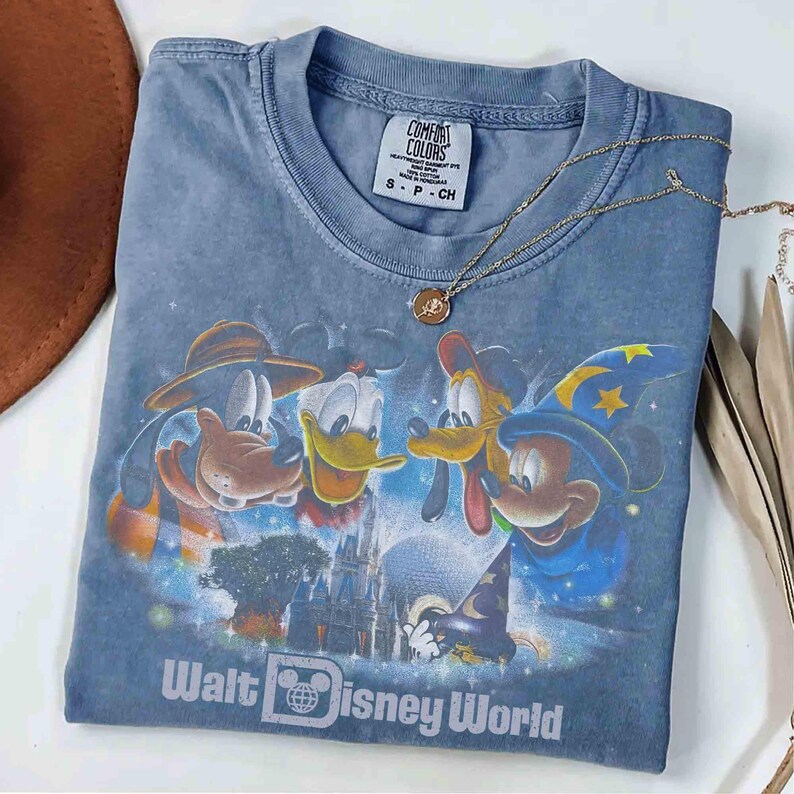 K&ouml;nnte beinhalten: Ein blaues Comfort Colors T-Shirt mit einem Aufdruck von Mickey Mouse, Goofy, Donald Duck und Pluto vor dem Cinderella-Schloss. Auf dem Shirt steht "Walt Disney World". Eine goldene Halskette liegt &uuml;ber dem Shirt.