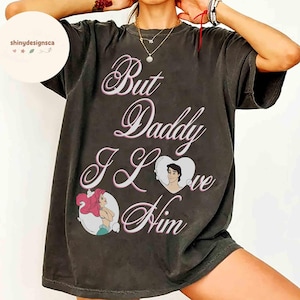 May include: Dark grey t-shirt with the text "But Daddy I Love Him" in a cursive font. The shirt features illustrations of Ariel and Prince Eric from The Little Mermaid. The shirt has a vintage, worn appearance.