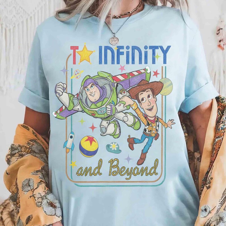 May include: A light blue t-shirt featuring a graphic print of Buzz Lightyear and Woody from Toy Story. The design includes the text "To INFINITY and Beyond" in blue and yellow, surrounded by stars, a rocket, a ball, and a hat inside a frame.