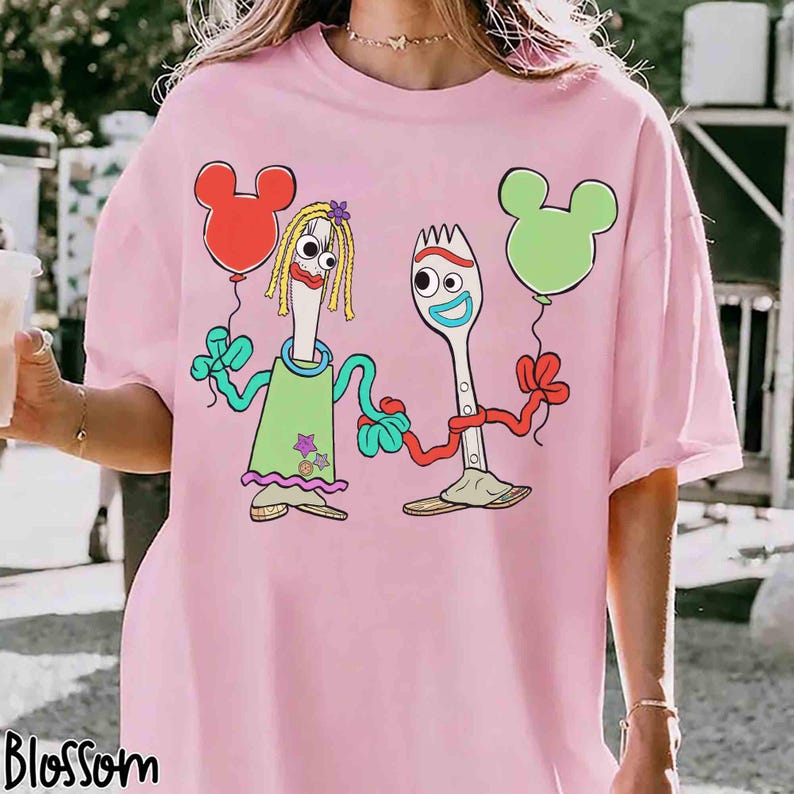 May include: Pink t-shirt featuring a cartoon illustration of two utensil characters holding Mickey Mouse-shaped balloons. One character is a fork wearing a green dress, and the other is a spoon. The word "Blossom" is printed at the bottom.
