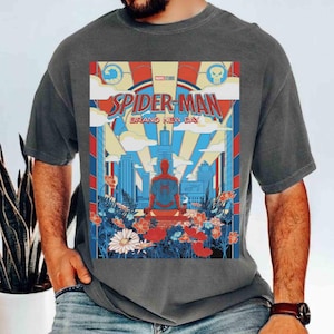May include: A charcoal gray t-shirt featuring a graphic print of Spider-Man sitting in front of a city skyline. The design includes the text "SPIDER-MAN GRAND NEW DAY" with red, blue, and yellow colors, surrounded by a field of colorful flowers.