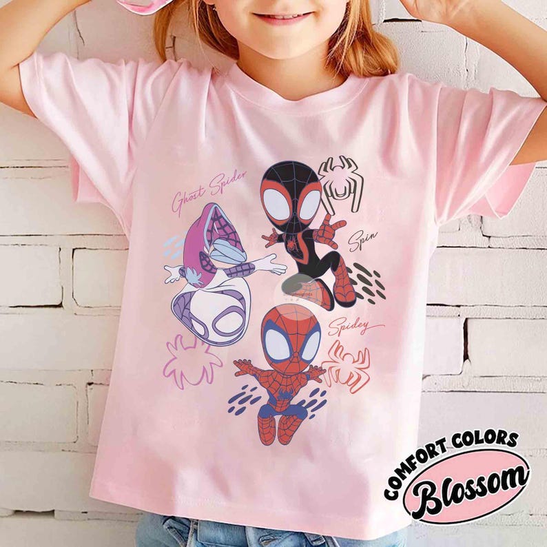 Op de afbeelding: Lichtroze T-shirt met cartoon illustraties van Spider-Man personages, waaronder Ghost Spider, Spin en Spidey. Het shirt heeft de woorden "Comfort Colors" en "Blossom" in een cirkelvormig ontwerp.