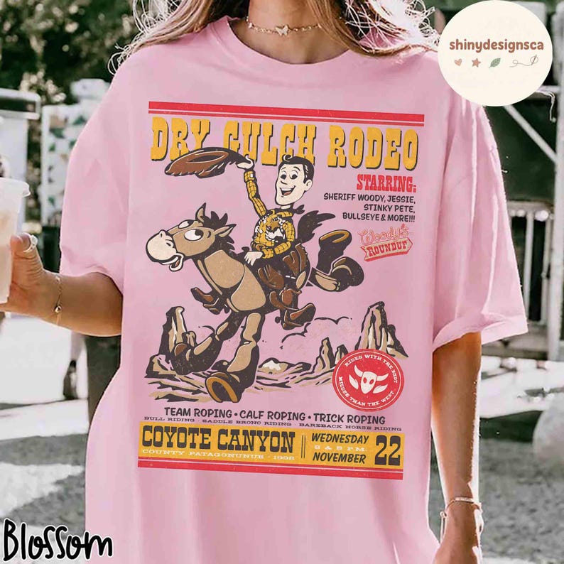 Puede incluir: Camiseta rosa claro con un dise&ntilde;o de rodeo de dibujos animados. El gr&aacute;fico incluye un vaquero a caballo, las palabras "Dry Gulch Rodeo" y "Coyote Canyon" con una fecha. El dise&ntilde;o tambi&eacute;n presenta la palabra "Starring" y nombres de personajes.