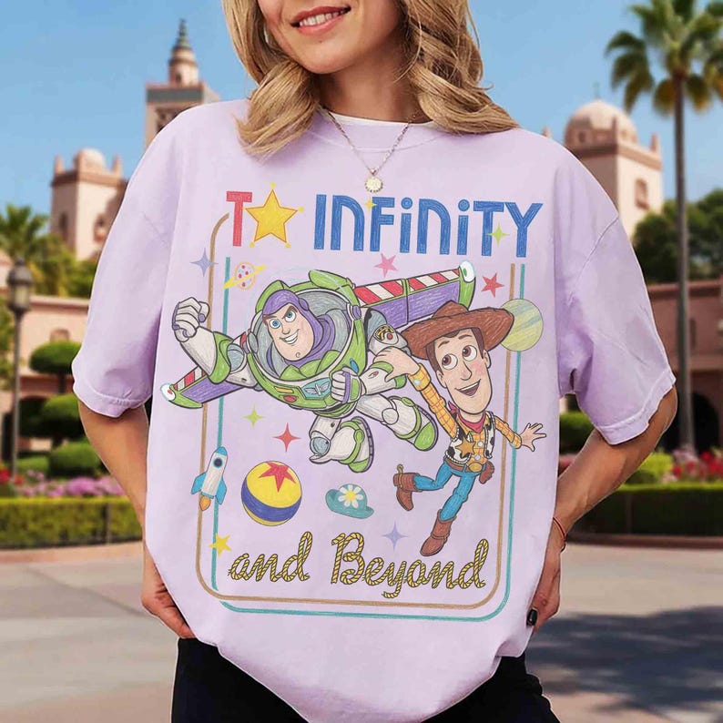 May include: A light purple t-shirt featuring a graphic print of Buzz Lightyear and Woody from Toy Story. The design includes the text "TO INFINITY and Beyond" in blue and yellow, surrounded by stars, a rocket, a ball, and a planet.