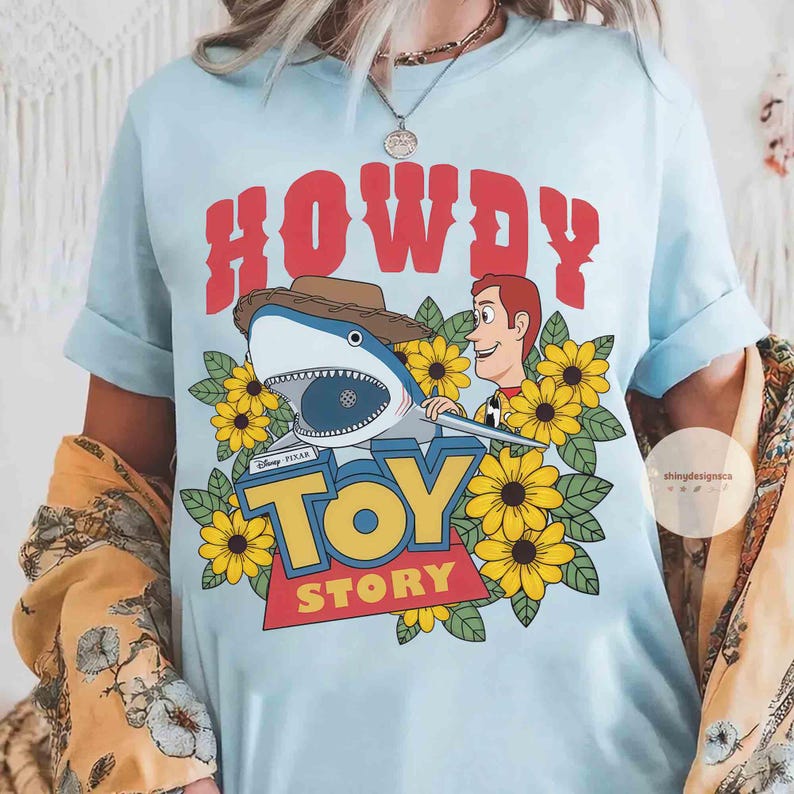 Puede incluir: Camiseta azul claro con un dise&ntilde;o gr&aacute;fico de la pel&iacute;cula de Disney Pixar Toy Story. El dise&ntilde;o incluye la palabra "HOWDY" en rojo, un tibur&oacute;n con sombrero de vaquero, Woody, girasoles amarillos y el logo de "TOY STORY".