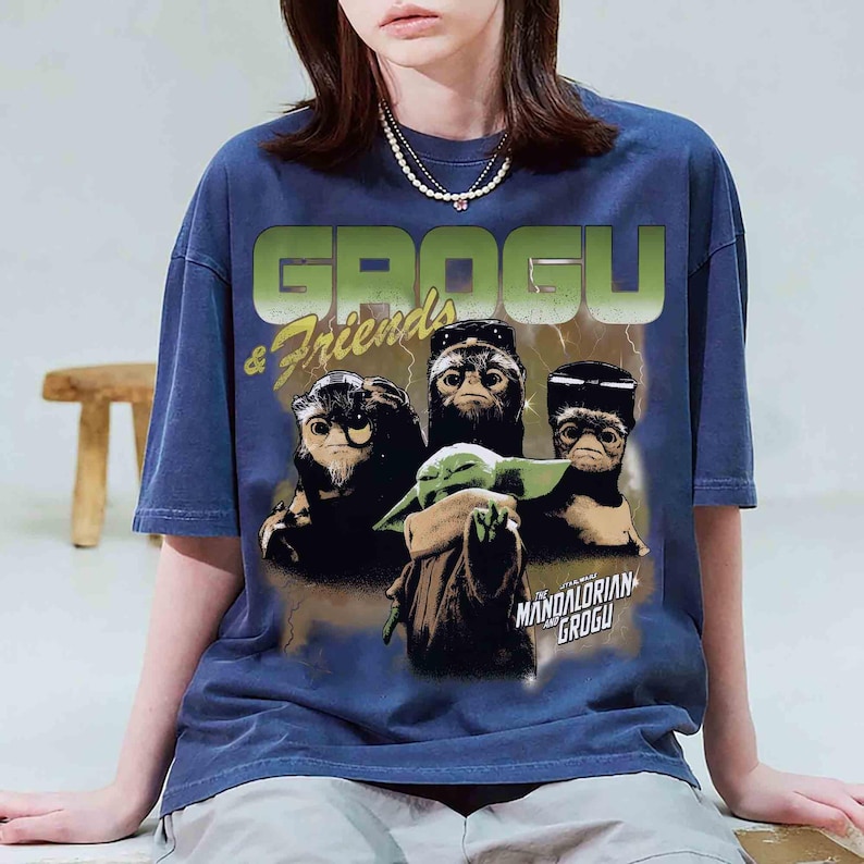 May include: Navy blue t-shirt featuring a graphic of Grogu and friends from The Mandalorian. The design includes the text "GROGU & Friends" in green and the phrase "The Mandalorian Grogu" in white. The shirt has short sleeves.