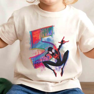 May include: Off-white t-shirt with a colourful graphic of the number 5 and a Spider-Man character. The Spider-Man is in a dynamic pose, with red and black accents. The graphic has a vibrant, comic book style.