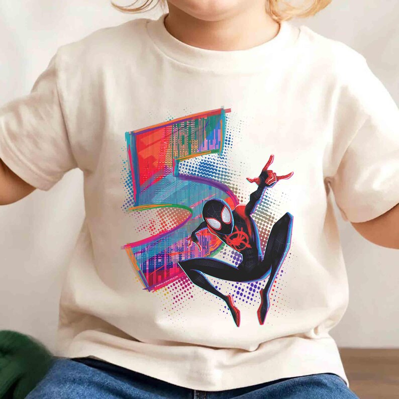 May include: Off-white t-shirt with a colourful graphic of the number 5 and a Spider-Man character. The Spider-Man is in a dynamic pose, with red and black accents. The graphic has a vibrant, comic book style.