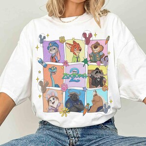 May include: White t-shirt featuring a colorful grid of Zootopia characters. The design includes Judy Hopps, Nick Wilde, and other characters from the animated film. The text "Zootopia 2" is also visible. The shirt has short sleeves.