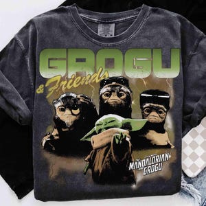 May include: Dark gray t-shirt featuring Grogu and other creatures from The Mandalorian. The shirt has the words "GROGU & Friends" in green and yellow, with a graphic of Grogu and other characters. The Star Wars logo is also visible.