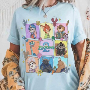 May include: Light blue t-shirt featuring a colorful graphic of characters from the animated movie Zootopia. The design includes Judy Hopps, Nick Wilde, and other characters in a grid pattern. The word "Zootopia" is also visible.