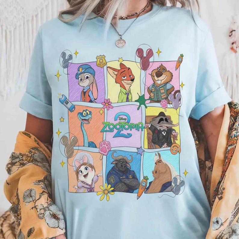 May include: Light blue t-shirt featuring a colorful graphic of characters from the animated movie Zootopia. The design includes Judy Hopps, Nick Wilde, and other characters in a grid pattern. The word "Zootopia" is also visible.