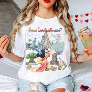 Op de afbeelding: Wit T-shirt met een kleurrijke afbeelding van Mickey Mouse als tovenaar, met de tekst "Some Imagination, Huh?" omringd door iconische Disney parkbeelden. Het ontwerp bevat een kasteel, een tovenaars hoed en andere parkelementen.