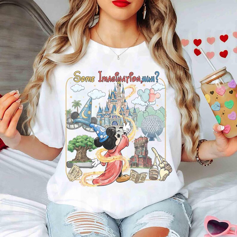 Op de afbeelding: Wit T-shirt met een kleurrijke afbeelding van Mickey Mouse als tovenaar, met de tekst "Some Imagination, Huh?" omringd door iconische Disney parkbeelden. Het ontwerp bevat een kasteel, een tovenaars hoed en andere parkelementen.