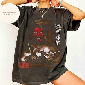 May include: Dark gray t-shirt featuring a graphic of a red-faced character and figures with lightsabers. Japanese text is printed on the shirt. The shirt has a vintage, worn look.