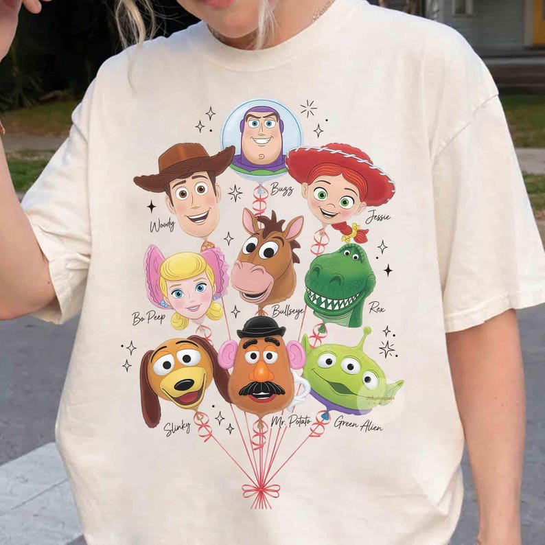 Op de afbeelding: Een cr&egrave;mekleurig t-shirt met een afbeelding van Toy Story-personages als ballonnen. Het ontwerp bevat Woody, Buzz, Jessie, Bo Peep, Bullseye, Rex, Slinky, Mr. Potato Head en Green Alien, elk met hun naam in cursief schrift gedrukt.