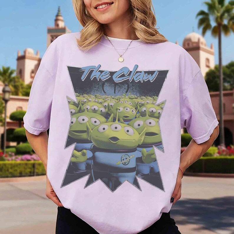 K&ouml;nnte beinhalten: Helllila T-Shirt mit einer Grafik von gr&uuml;nen, dre&auml;ugigen Aliens in blauen Uniformen. Der Text "The Claw" steht in blauer Schrift &uuml;ber den Aliens, innerhalb eines gezackten schwarzen und grauen Rahmens.