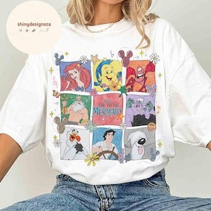 May include: White t-shirt featuring a colorful graphic of characters from Disney's "The Little Mermaid." The design includes Ariel, Sebastian, Flounder, Ursula, and other characters in a grid pattern. The shirt has a crew neck and short sleeves.