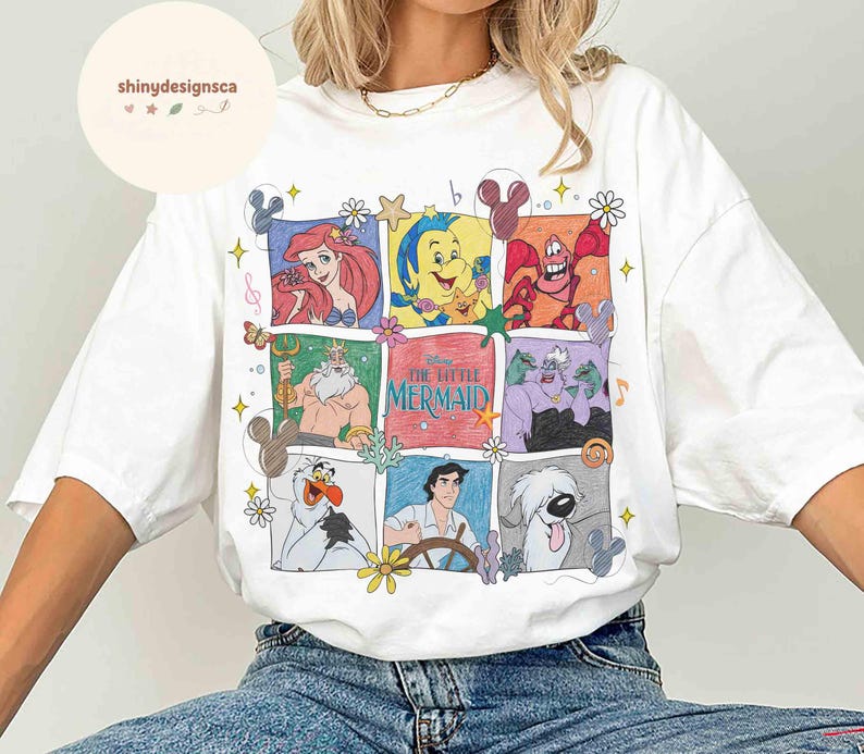 May include: White t-shirt featuring a colorful graphic of characters from Disney's "The Little Mermaid." The design includes Ariel, Sebastian, Flounder, Ursula, and other characters in a grid pattern. The shirt has a crew neck and short sleeves.