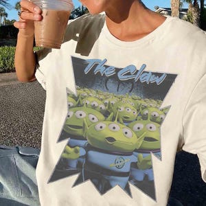 K&ouml;nnte beinhalten: Cremefarbenes T-Shirt mit einer Grafik von gr&uuml;nen, dre&auml;ugigen Aliens in blauen Uniformen und dem Text "The Claw" in Hellblau. Die Aliens sind vor einem dunkelblauen Hintergrund in einer gezackten schwarz-wei&szlig;en Form platziert.