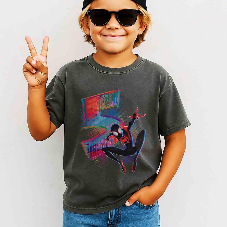 May include: Dark grey t-shirt with a colourful Spider-Man graphic and the number 5. The design includes a stylized cityscape and vibrant colour accents. The shirt is worn by a child.