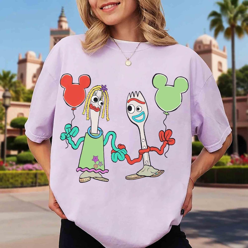 May include: A light purple t-shirt featuring a cartoon illustration of two characters holding Mickey Mouse-shaped balloons. One character is a fork with a blue face and the other is a fork with blonde hair. The shirt has a relaxed fit.