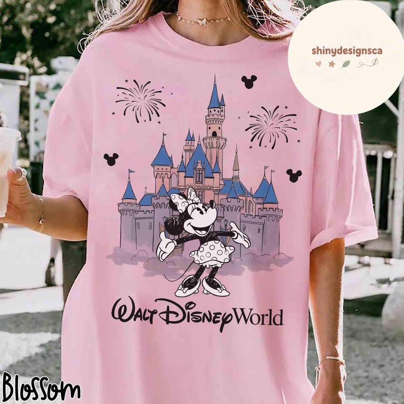 May include: Pink t-shirt featuring a Minnie Mouse graphic in front of a castle with blue turrets and fireworks. The words "Walt Disney World" are printed below the graphic. The shirt has a round neck and short sleeves.