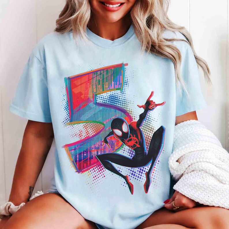 May include: Pale blue t-shirt with a colourful graphic of the number 5 and a Spider-Man character in a dynamic pose. The design uses vibrant red, blue, and purple hues, creating a striking visual.