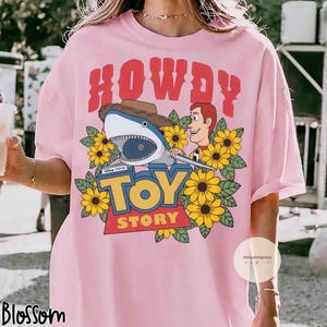 Puede incluir: Camiseta rosa claro con un dise&ntilde;o de la pel&iacute;cula Toy Story. El dise&ntilde;o incluye la palabra "HOWDY" en rojo, un tibur&oacute;n con sombrero de vaquero, Woody, girasoles y las palabras "TOY STORY".