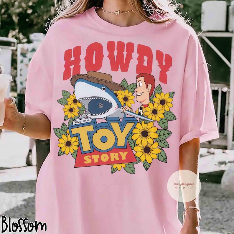 Puede incluir: Camiseta rosa claro con un dise&ntilde;o de la pel&iacute;cula Toy Story. El dise&ntilde;o incluye la palabra "HOWDY" en rojo, un tibur&oacute;n con sombrero de vaquero, Woody, girasoles y las palabras "TOY STORY".