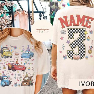 以下が含まれることがあります： アイボリー色のTシャツ。漫画の車、カップケーキ、誕生日の装飾がカラフルに描かれています。Tシャツの背面には、市松模様の数字「3」の上に「NAME」の文字と、誕生日のイラストが描かれています。