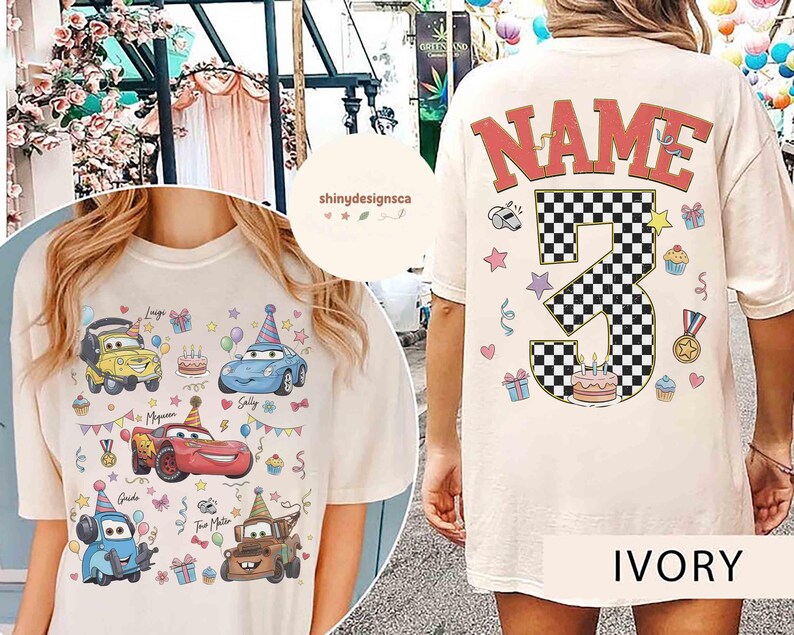 以下が含まれることがあります： アイボリー色のTシャツ。漫画の車、カップケーキ、誕生日の装飾がカラフルに描かれています。Tシャツの背面には、市松模様の数字「3」の上に「NAME」の文字と、誕生日のイラストが描かれています。