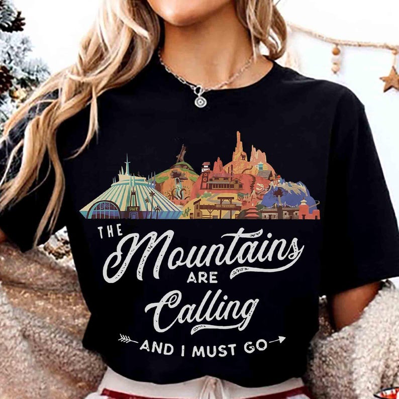May include: A black short-sleeve t-shirt featuring a colorful graphic of various mountain-themed attractions. The text reads "THE Mountains ARE Calling AND I MUST GO" in white script and block lettering, with a small arrow pointing to the right.