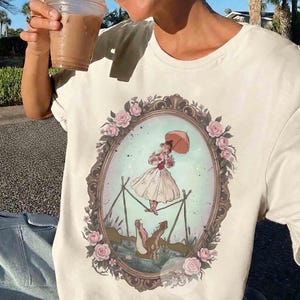 Puede incluir: Una camiseta de color crema con un estampado gr&aacute;fico de un marco ovalado ornamentado decorado con rosas rosas. Dentro del marco, una figura con vestido blanco sostiene una sombrilla mientras camina sobre una cuerda floja sobre un caim&aacute;n.