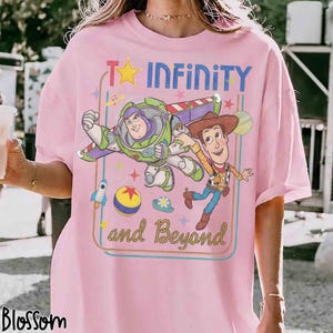 May include: A light pink t-shirt featuring a graphic print of Buzz Lightyear and Woody from Toy Story. The design includes the text "TO INFINITY and Beyond", a yellow star, a rocket, a ball, and various colorful stars on a light pink background.