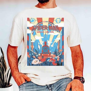 May include: A cream-colored t-shirt featuring a graphic print of Spider-Man sitting in a meditative pose. The design includes a city skyline, clouds, and colorful flowers in red, blue, and white, with the text Spider-Man Brand New Day.